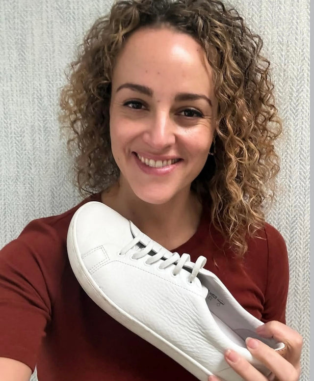 A smiling woman with curly hair holding up a well-worn white leather Birchbury sneaker against a textured grey wall