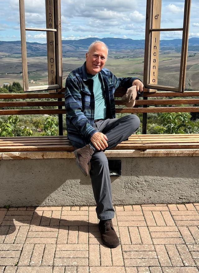 An older man in a blue plaid jacket smiling on a bench overlooking the Tuscan countryside, wearing brown suede Birchbury chukka boots