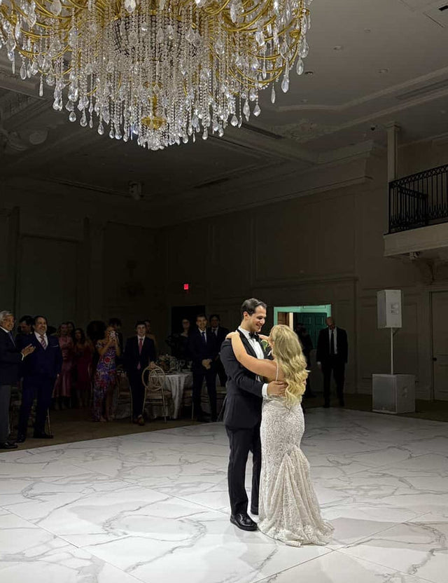 A groom in a black tuxedo and bride in a lace wedding gown share their first dance on a marble ballroom floor beneath an ornate crystal chandelier