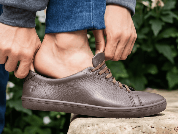 A person adjusting the heel of a brown leather Birchbury sneaker while standing on a stone ledge surrounded by white flowering shrubs