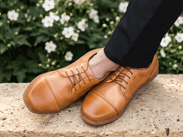 Overhead view of tan leather cap-toe Birchbury oxfords with crossed ankles, worn with black dress trousers on a stone-tiled sidewalk