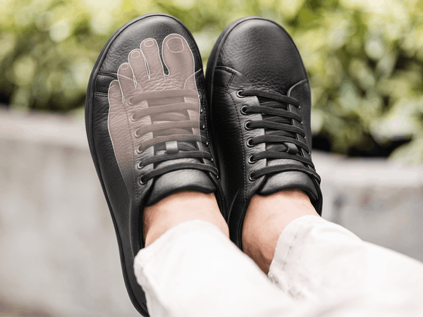 Black leather Birchbury sneakers with a translucent foot outline overlaid on one shoe, showing how the wide toe box gives toes room to spread naturally